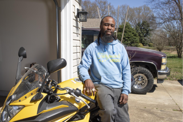 A photo of Demetre, a Self-Help member, leaning on a motorcycle in the driveway of his home