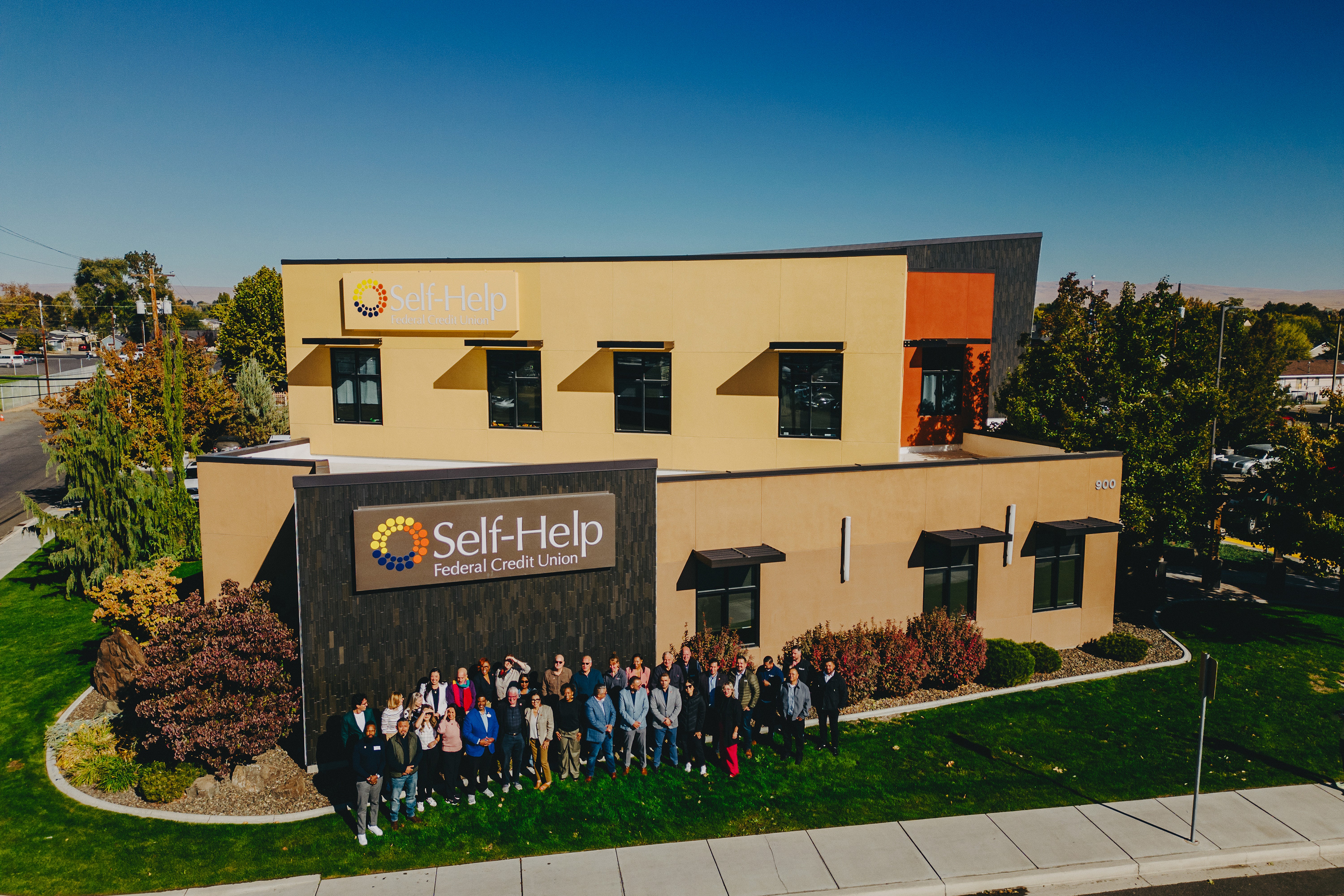 An aerial shot or perspective of a group of people standing on a lawn in front of a Self-Help Federal Credit Union building